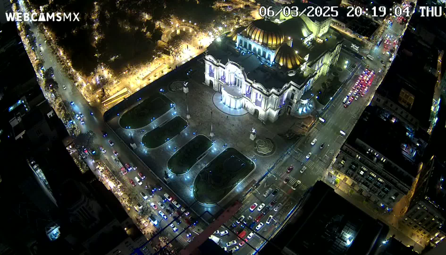 An illuminated palace-like building sits in a city square at night, surrounded by roadways with moderate traffic under clear skies.