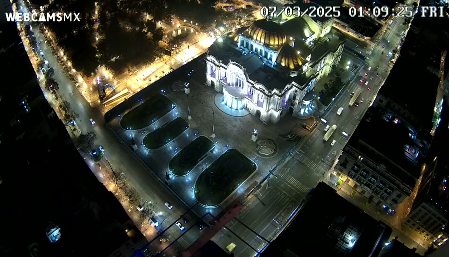 An illuminated Palacio de Bellas Artes at night, surrounded by city streets with cars and a plaza with landscaping.