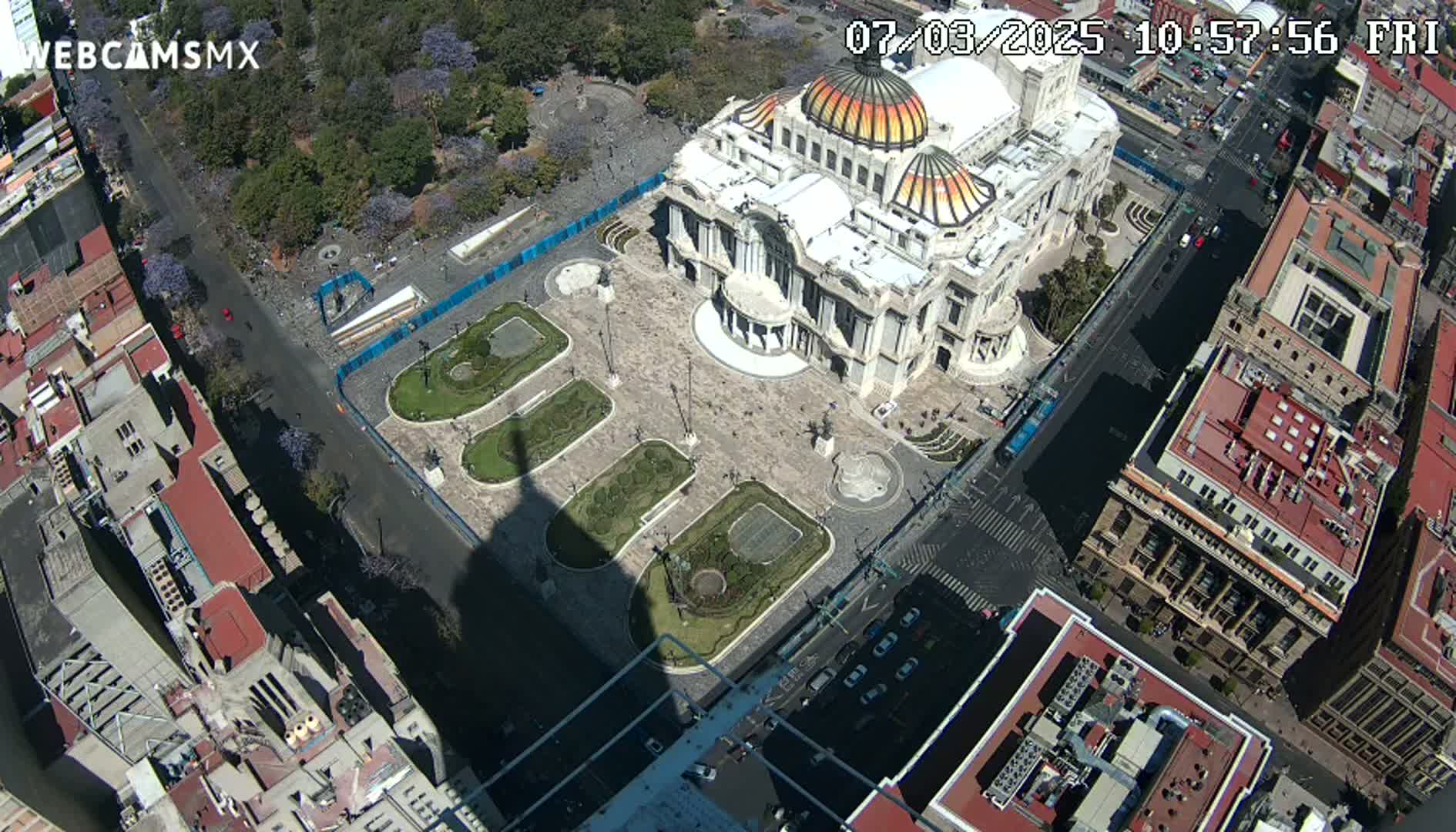 A high-angle view shows a large, ornate building in a plaza with surrounding city buildings under sunny conditions.
