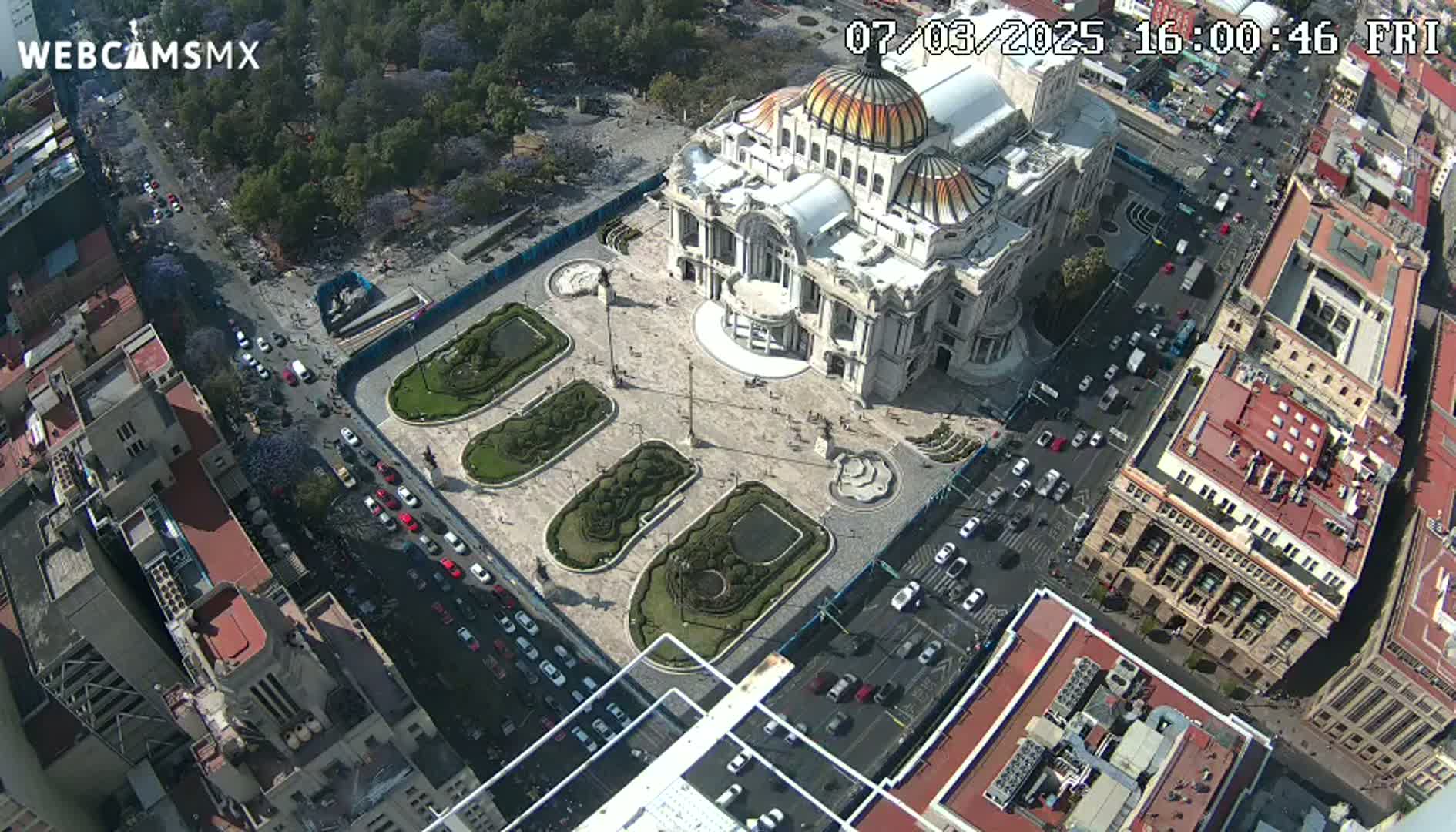 An aerial view shows a large building with a dome, surrounded by a plaza with landscaped gardens, and busy streets with cars on a sunny day.