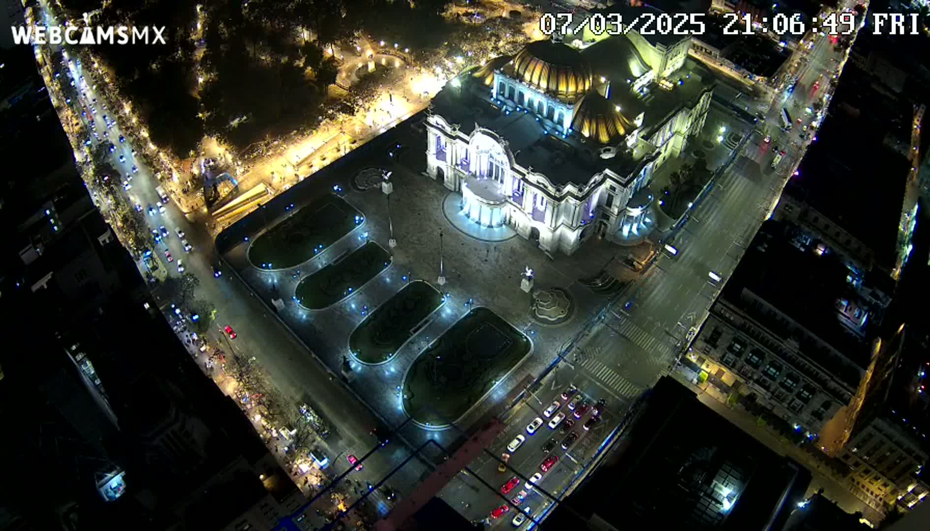An aerial nighttime view shows a large, ornate building illuminated, surrounded by streets with cars and pedestrians, and landscaped areas.