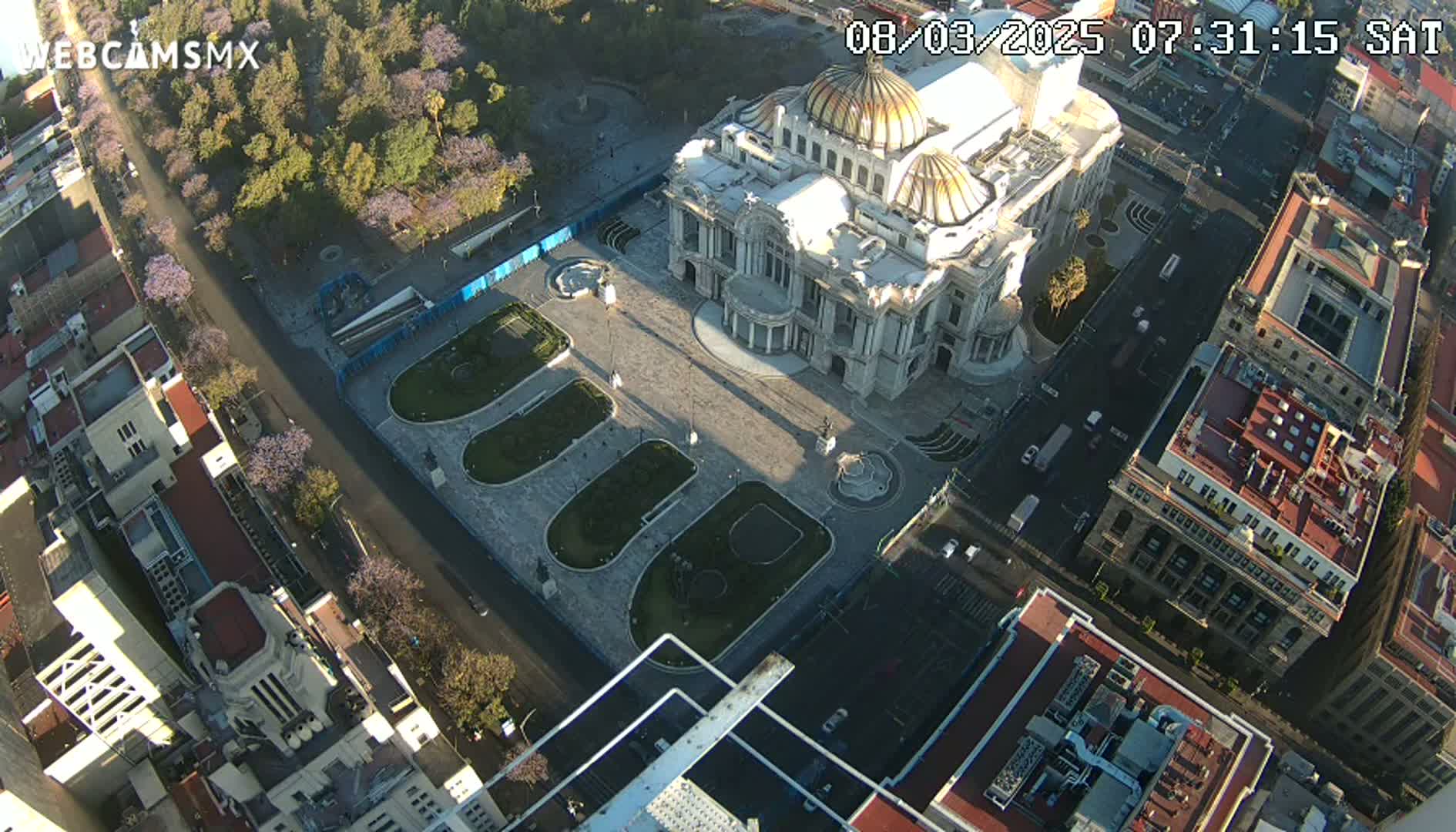 An aerial view shows a large, ornate building surrounded by a plaza with landscaped areas, city streets, and other buildings under a clear, sunny sky.