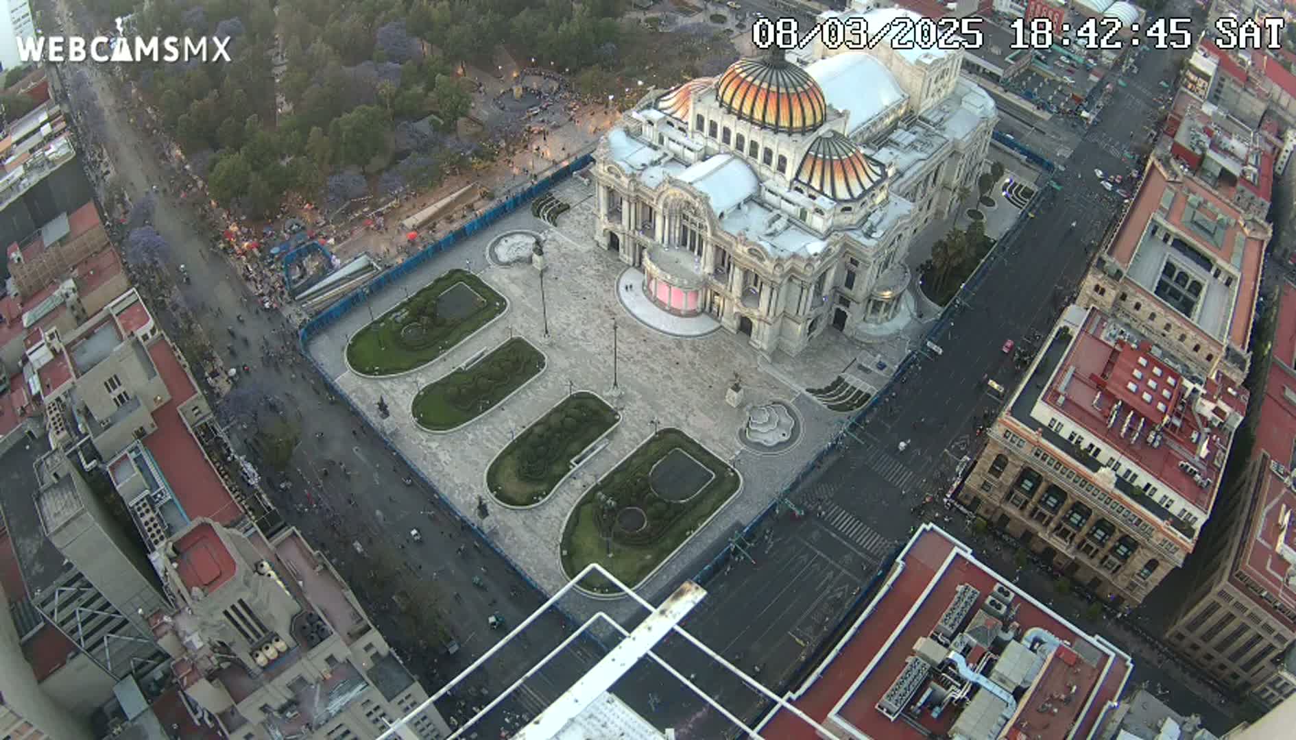 An aerial view shows a large building with a dome surrounded by a plaza with landscaped areas, all situated in a city with streets and other buildings visible, under what appears to be twilight conditions.