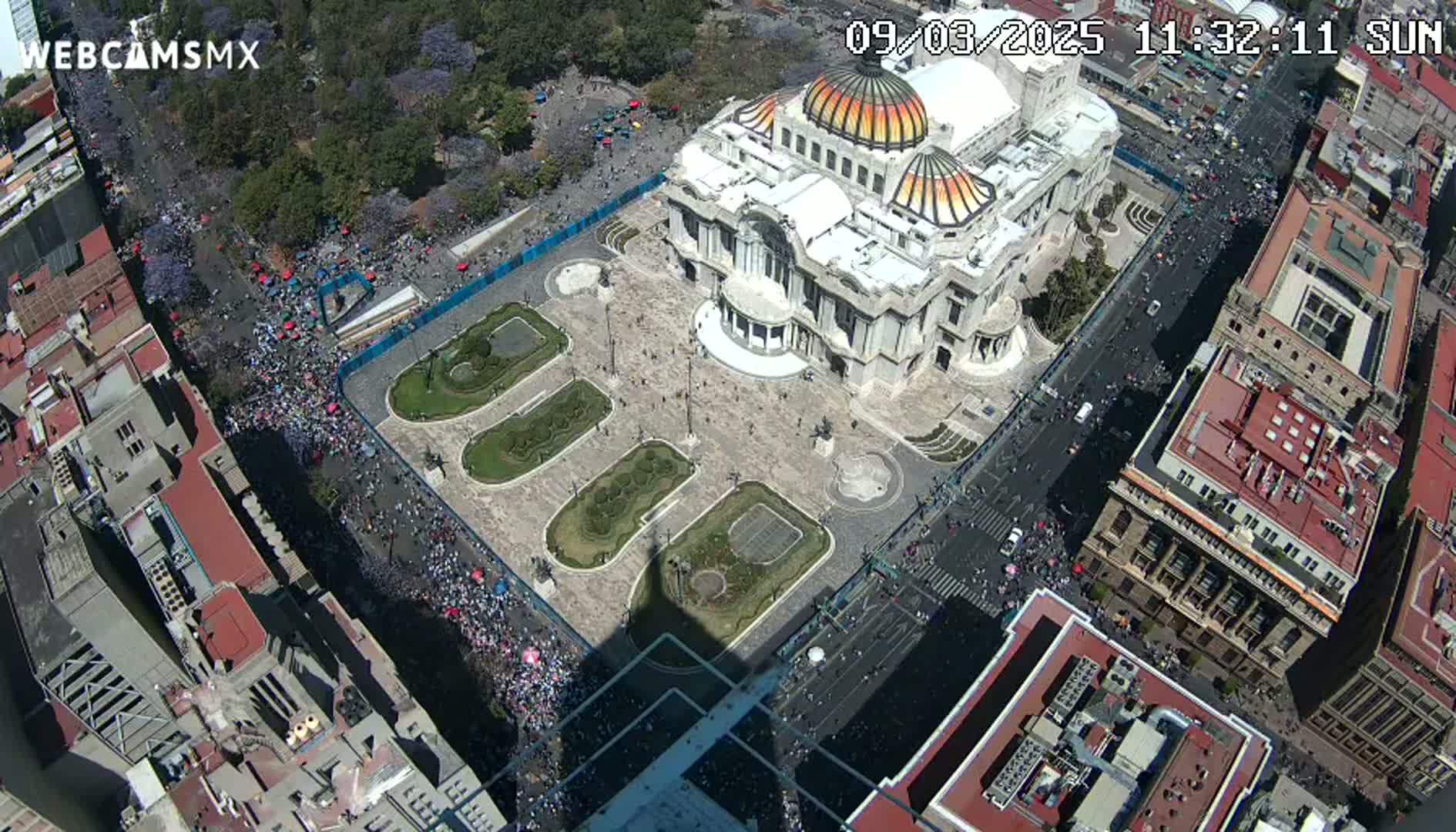 An aerial view shows a large crowd gathered in a city square surrounding a grand building on a sunny day.