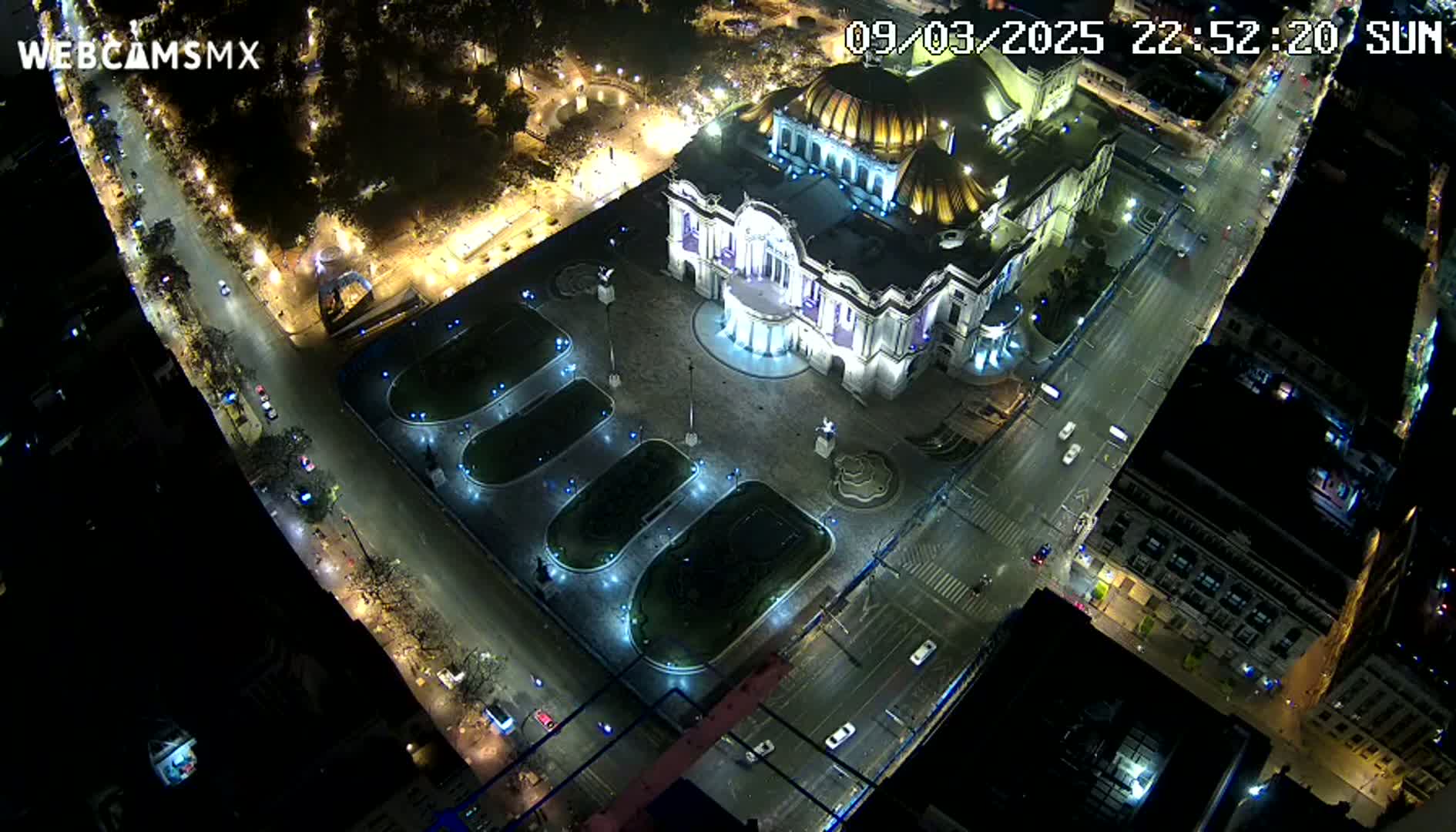 A brightly lit palace-like building sits in a plaza at night, surrounded by streets with sparse traffic under a clear sky.