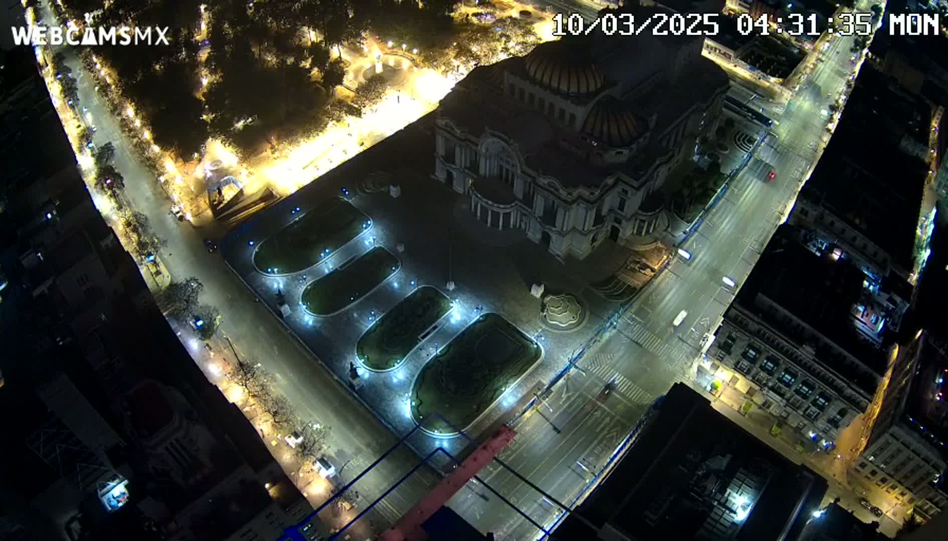 A nighttime aerial view shows a large building illuminated, surrounded by streets and a plaza with landscaping, under clear skies.