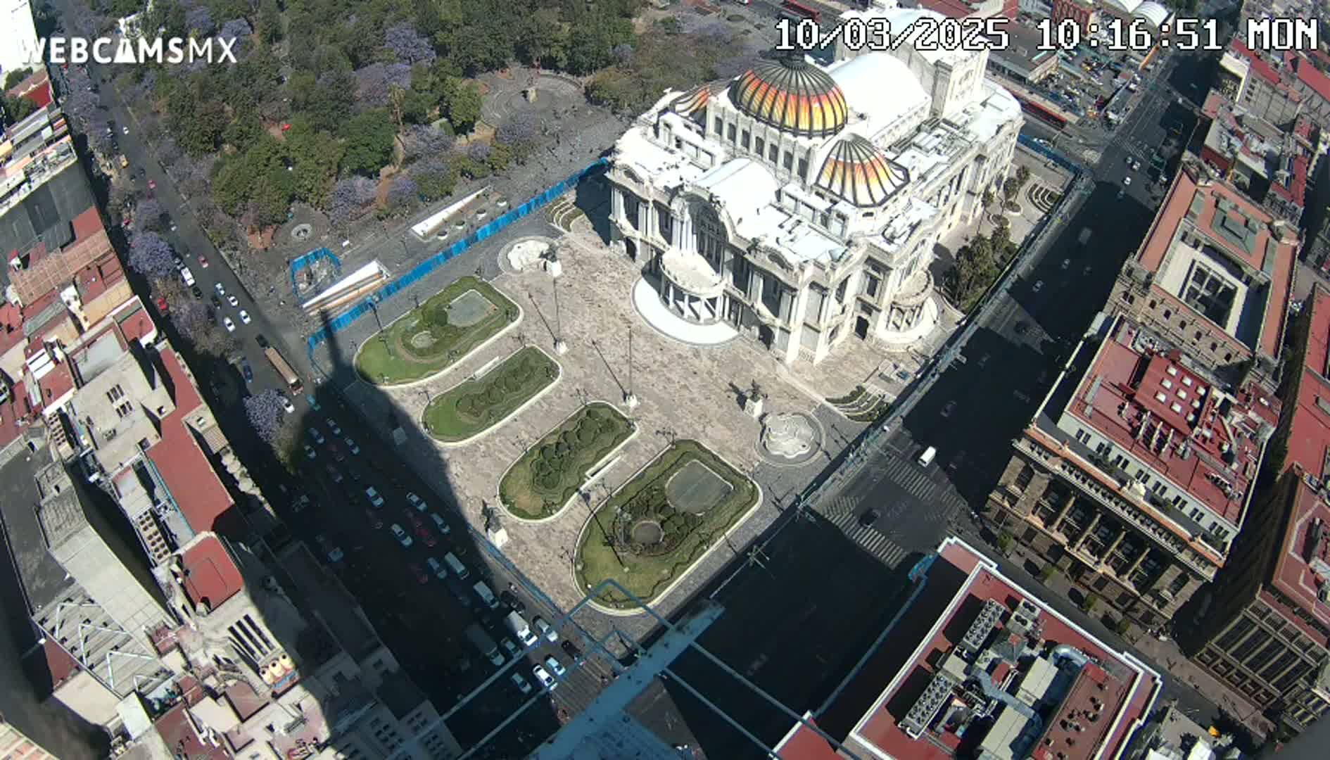 An aerial view shows a large building with a domed roof surrounded by a plaza with landscaped gardens and city streets with light traffic on a sunny day.