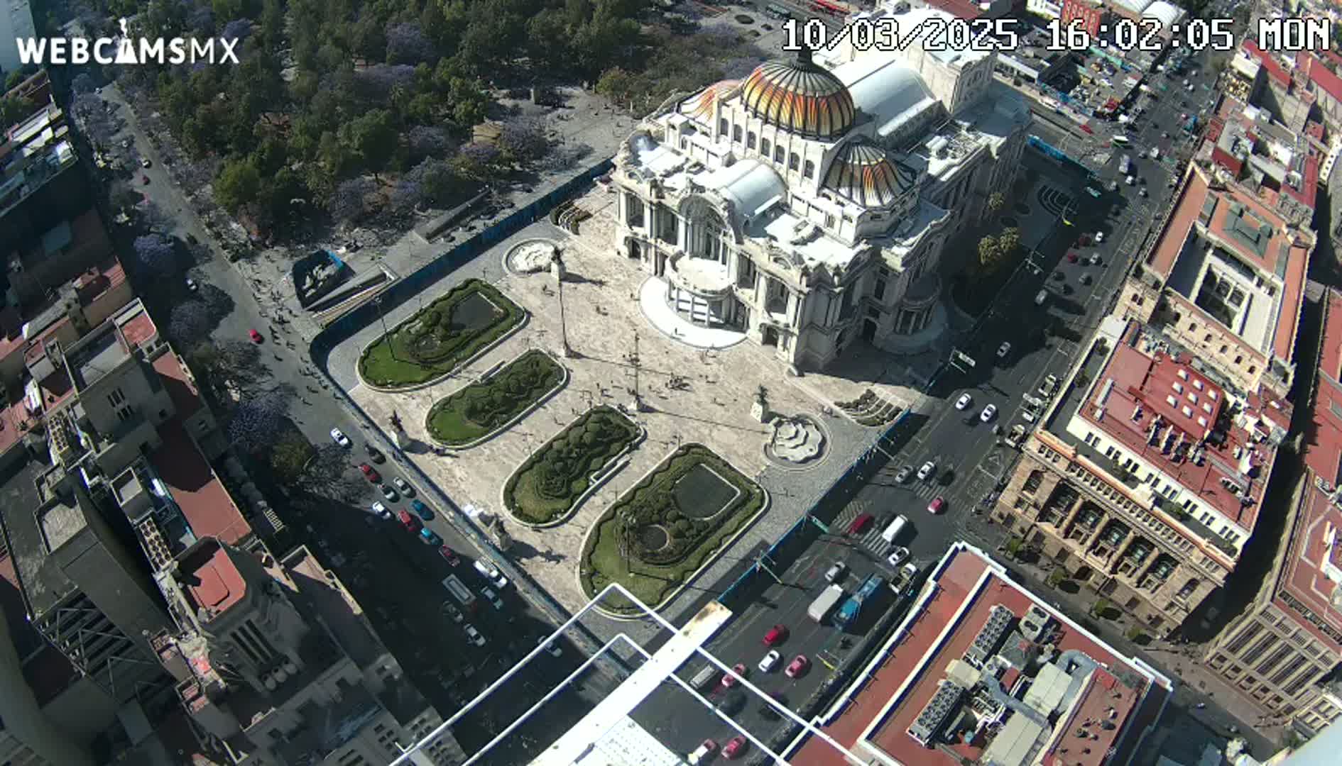 An aerial view shows a large, ornate building surrounded by a plaza with landscaped gardens and streets filled with cars under a sunny sky.