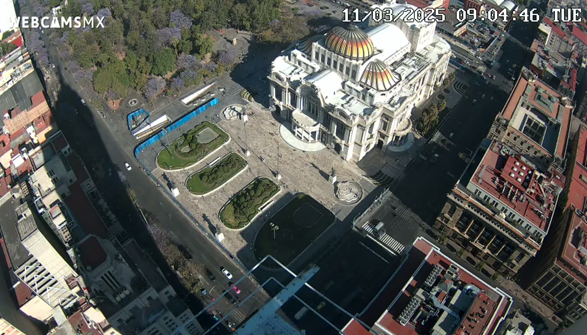 An aerial view shows a large, ornate building in a city square surrounded by streets and other buildings on a sunny day.