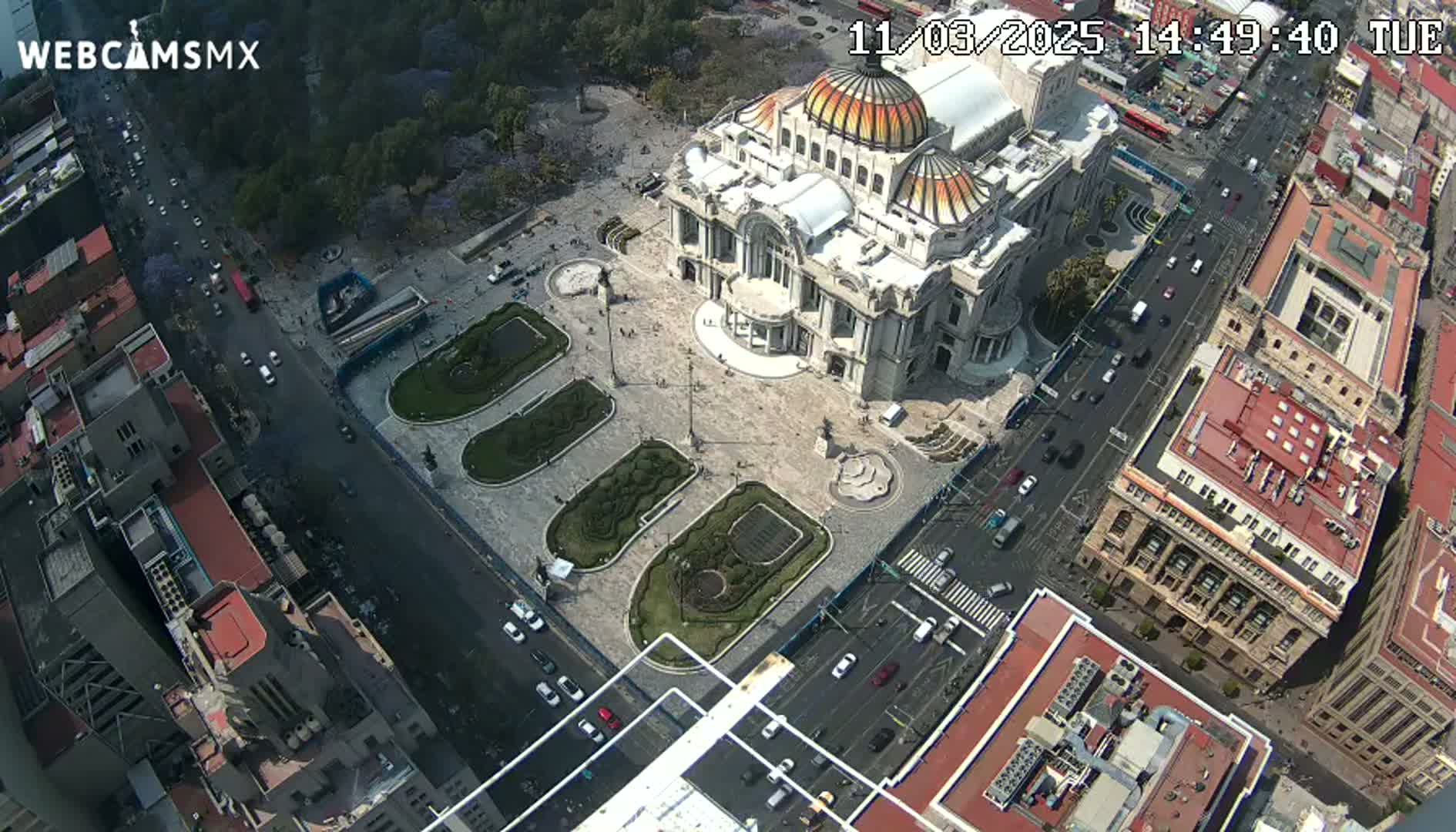 An aerial view shows a large building with a dome, surrounded by a plaza with landscaping and city streets with cars on a sunny day.