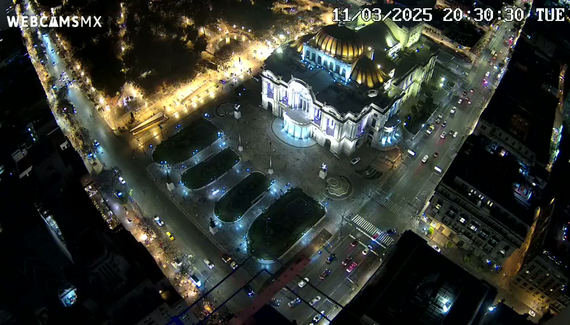 An aerial nighttime view shows a large, ornate building illuminated, surrounded by streets with cars and a plaza with landscaping, under clear night skies.