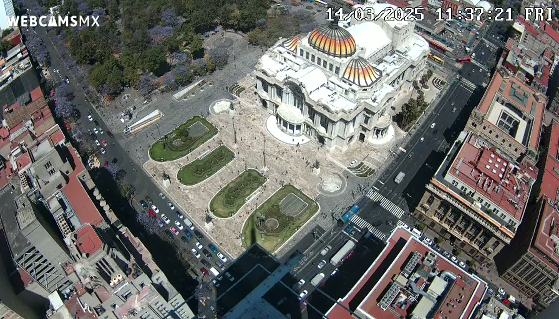 An aerial view shows a large building with a domed roof surrounded by a plaza with landscaped gardens and city streets with cars under a sunny sky.