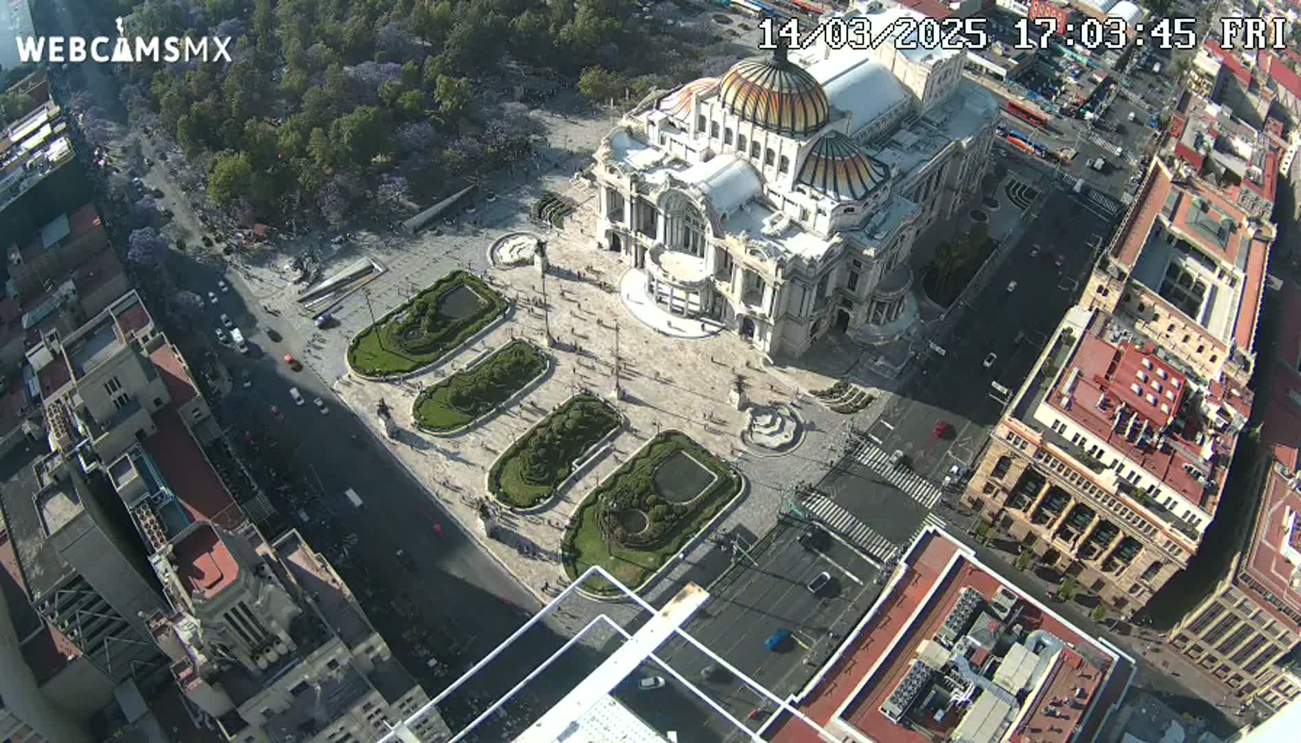 An aerial view shows a large plaza with a grand building and surrounding city structures on a sunny day.