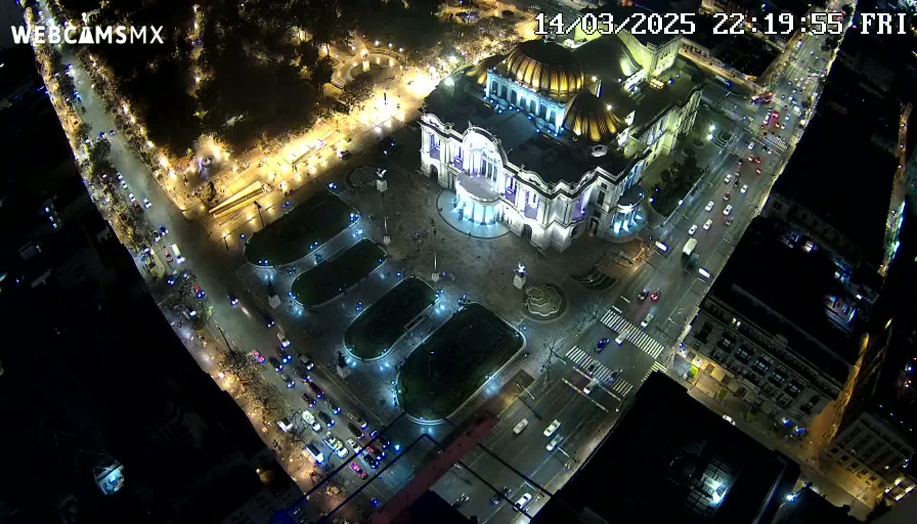 A nighttime aerial view shows a large, illuminated building surrounded by streets with cars, and a plaza with landscaped areas.