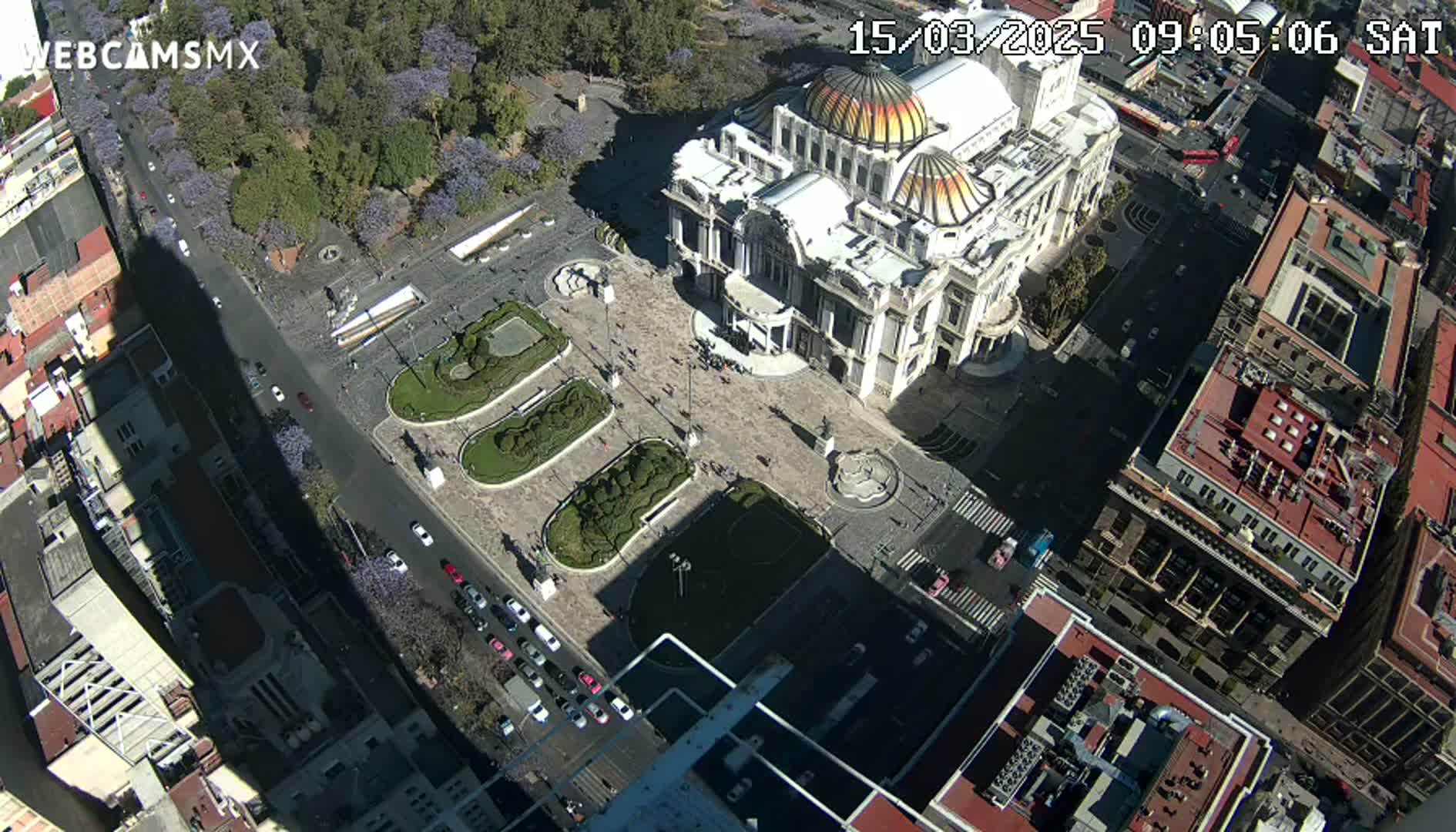 A high-angle, sunny view of a city square with a large building in the center, surrounded by streets and other buildings.