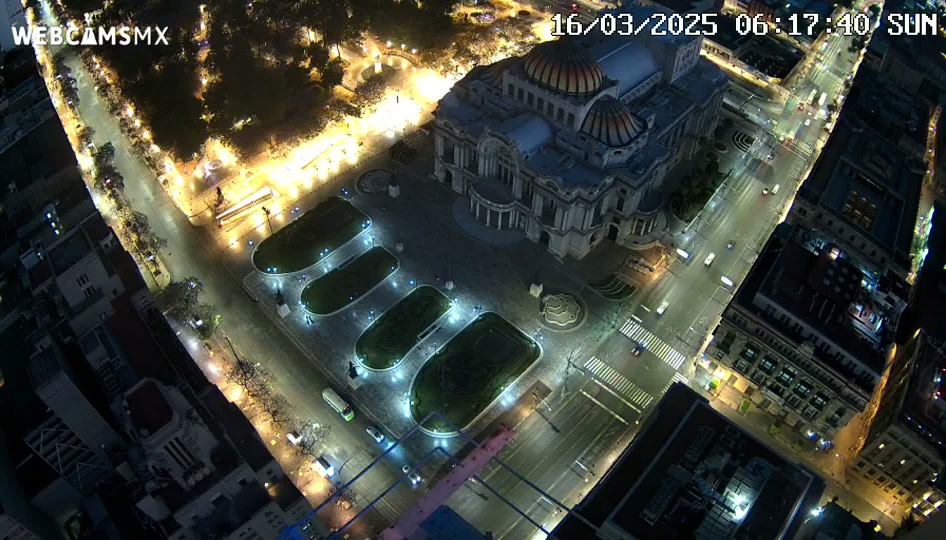 An aerial nighttime view of a city square with a large building, illuminated streets, and several vehicles.