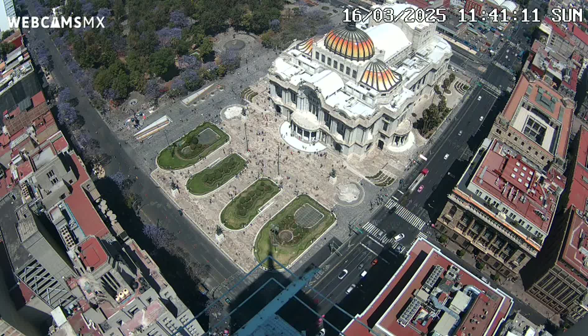 A high-angle, sunny view shows a large plaza with a grand building at its center, surrounded by city streets and buildings.
