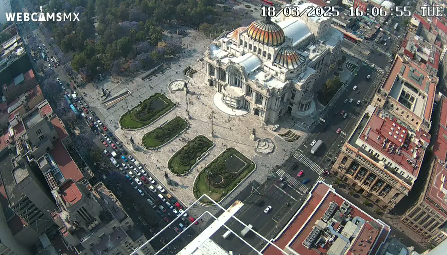 An aerial view shows a large plaza with a grand building at its center, surrounded by city streets and buildings under a sunny sky.