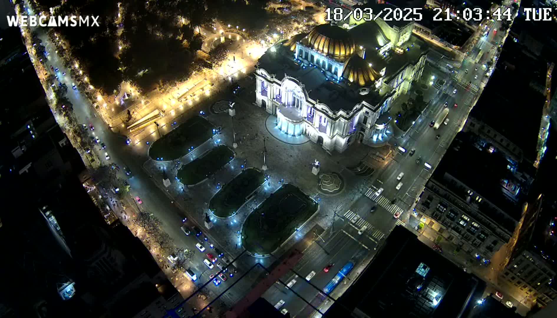 An aerial nighttime view shows a large, illuminated building in a city square with surrounding streets and vehicles, under clear skies.