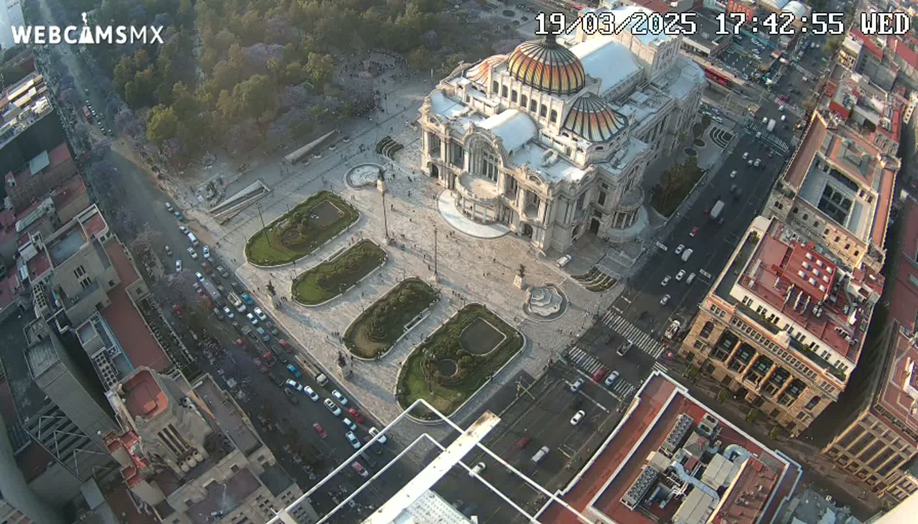 An aerial view shows a large, ornate building in a city square surrounded by streets with cars and other buildings under a partly sunny sky.
