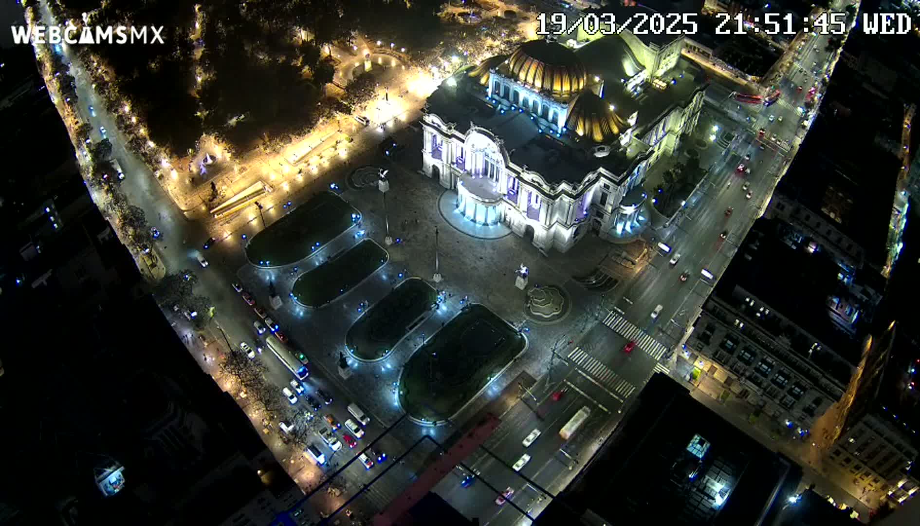 An aerial nighttime view shows a large, illuminated building in a city square with surrounding streets and traffic.