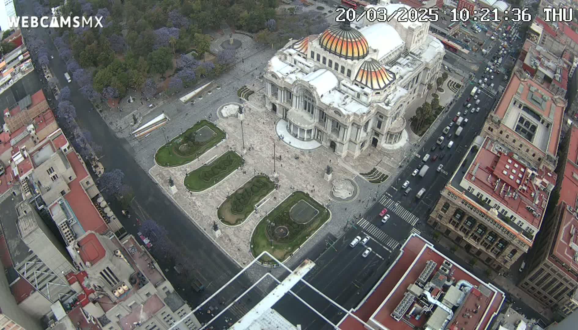 An aerial view of a large building with a dome, surrounded by a plaza with landscaped areas and city streets with cars under an overcast sky.