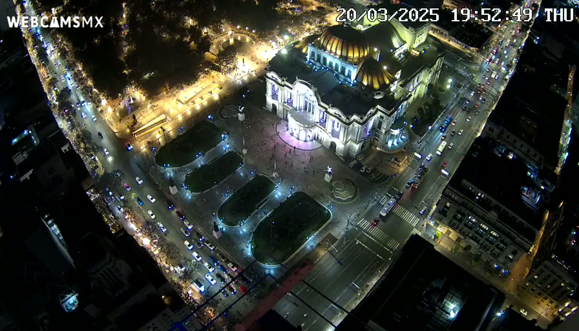 An aerial nighttime view shows a large building illuminated, surrounded by a plaza with landscaping and roads filled with vehicles.