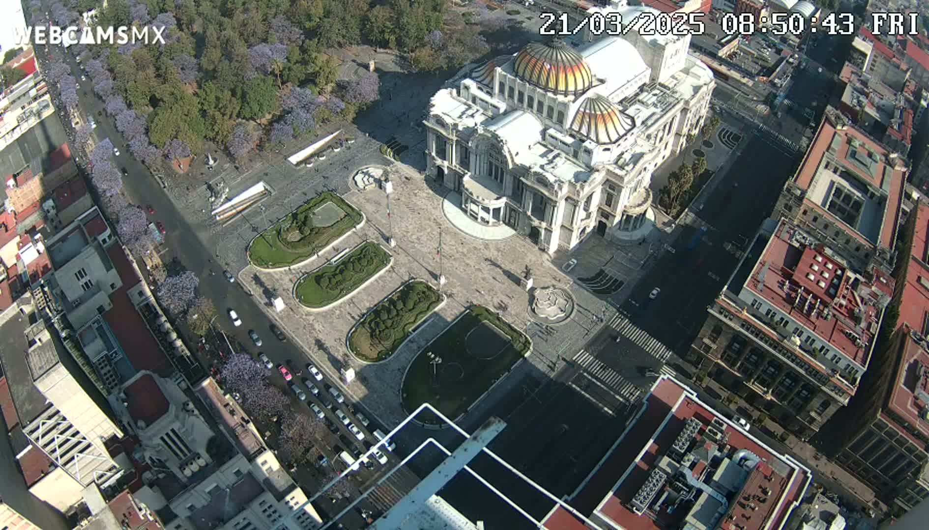 An aerial view shows a large, ornate building situated in a plaza with landscaped gardens, surrounded by city streets and buildings on a sunny day.