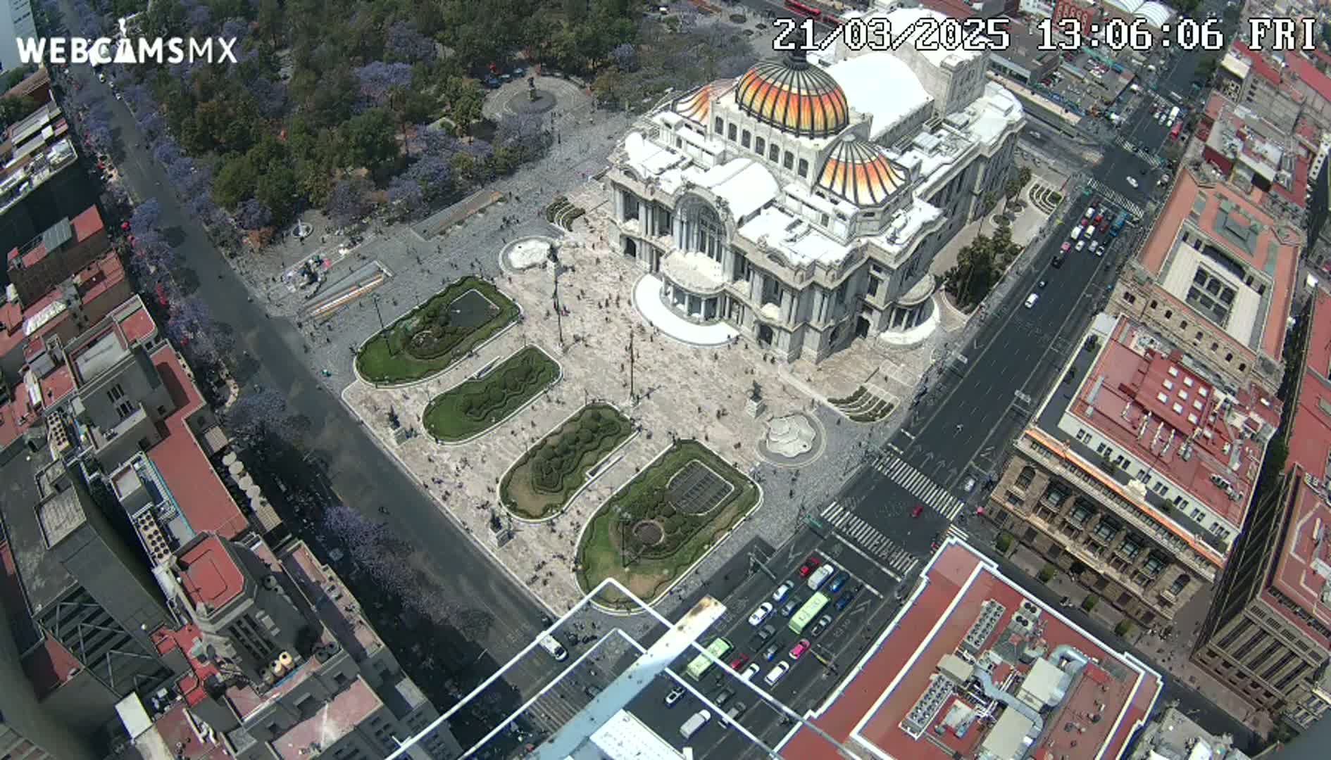 An aerial view shows a large, ornate building surrounded by a plaza with landscaped gardens and city streets with cars on a sunny day.