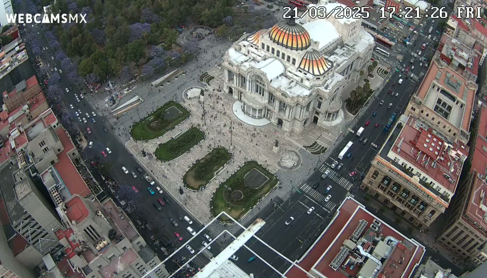 An aerial view shows a large plaza with a grand building at its center, surrounded by city streets with traffic and buildings under a mostly clear sky.