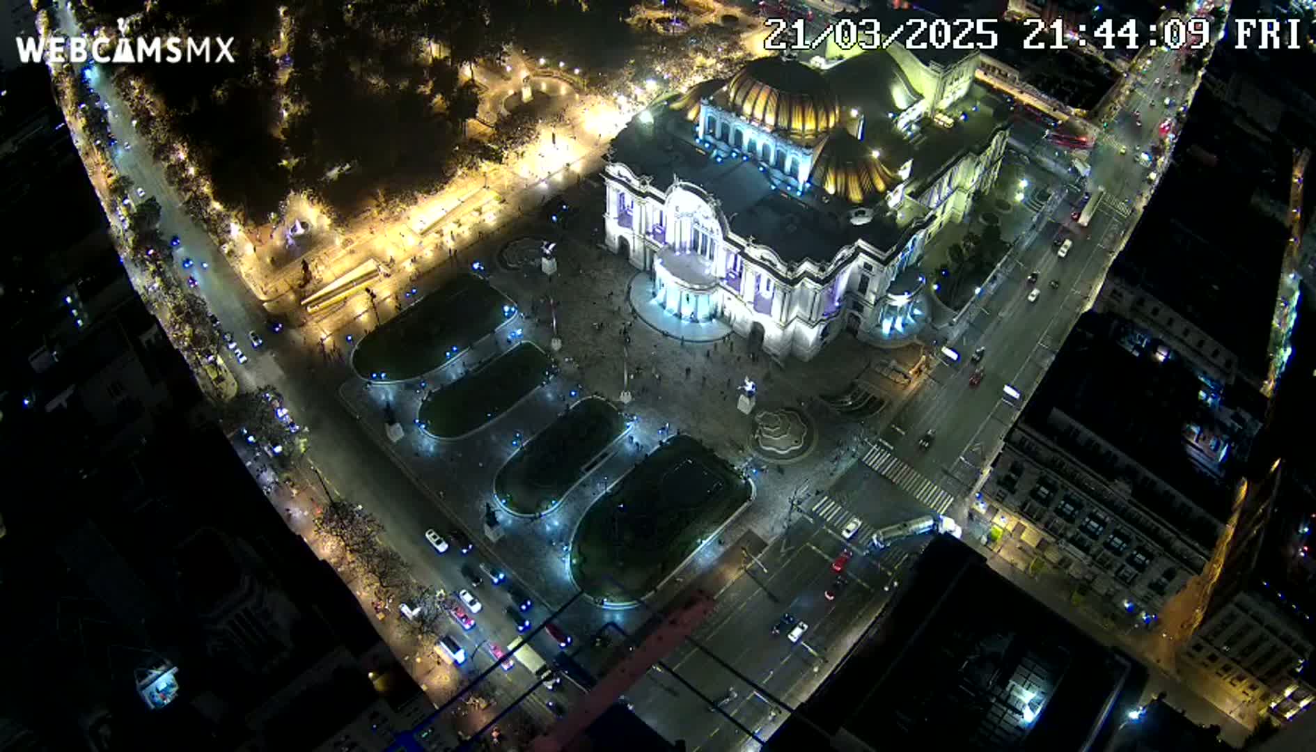 A nighttime aerial view shows a large, illuminated building surrounded by streets and a plaza with people and vehicles, under clear skies.