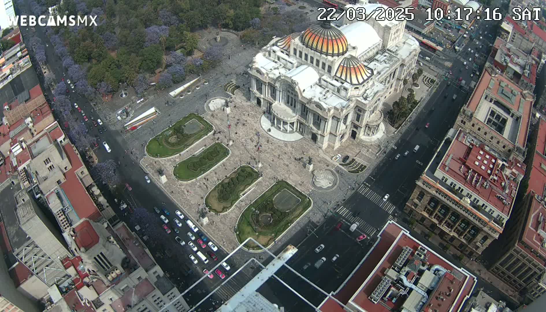 An aerial view shows a large, ornate building in a city square with surrounding streets and buildings on a sunny day.