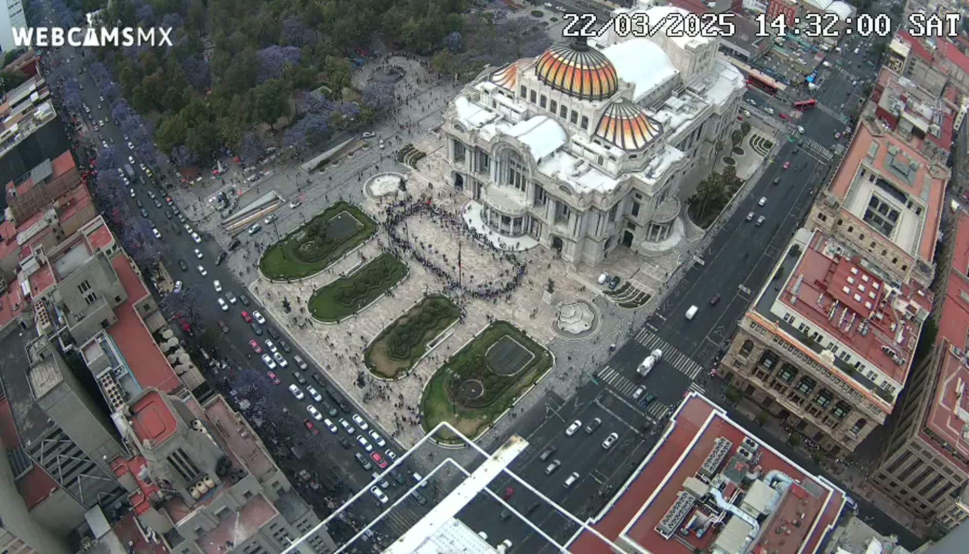 An aerial view shows a large building surrounded by a plaza with a crowd of people, streets with cars, and surrounding city buildings under a clear sky.