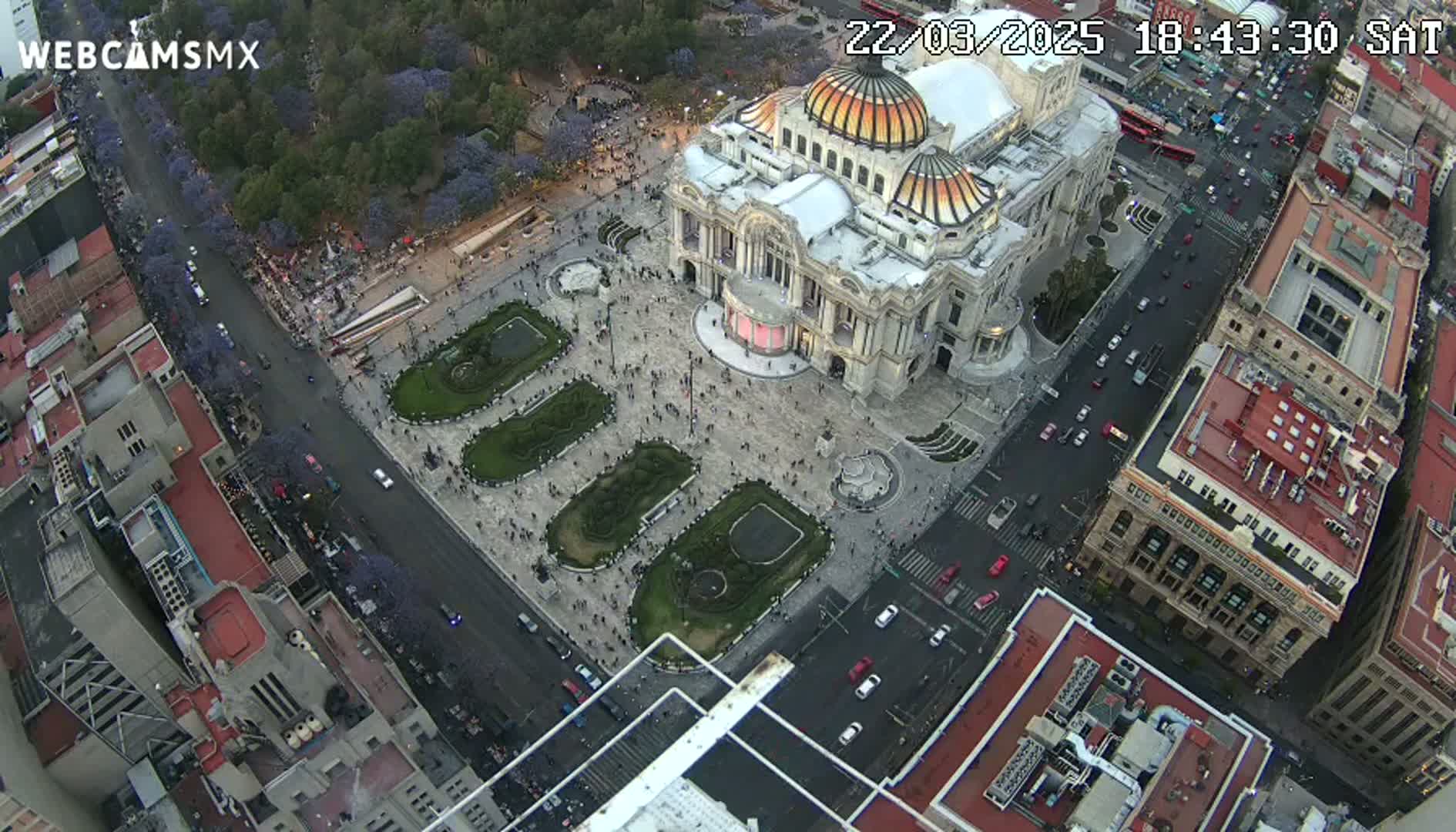 An aerial view shows a large building surrounded by a plaza with people and landscaping, all under a twilight sky.