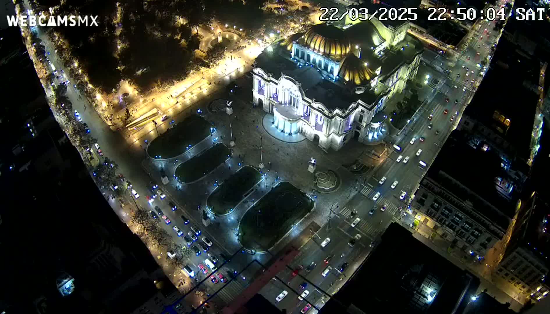 A nighttime aerial view shows a large, illuminated building surrounded by a plaza with cars driving on surrounding streets under a clear sky.