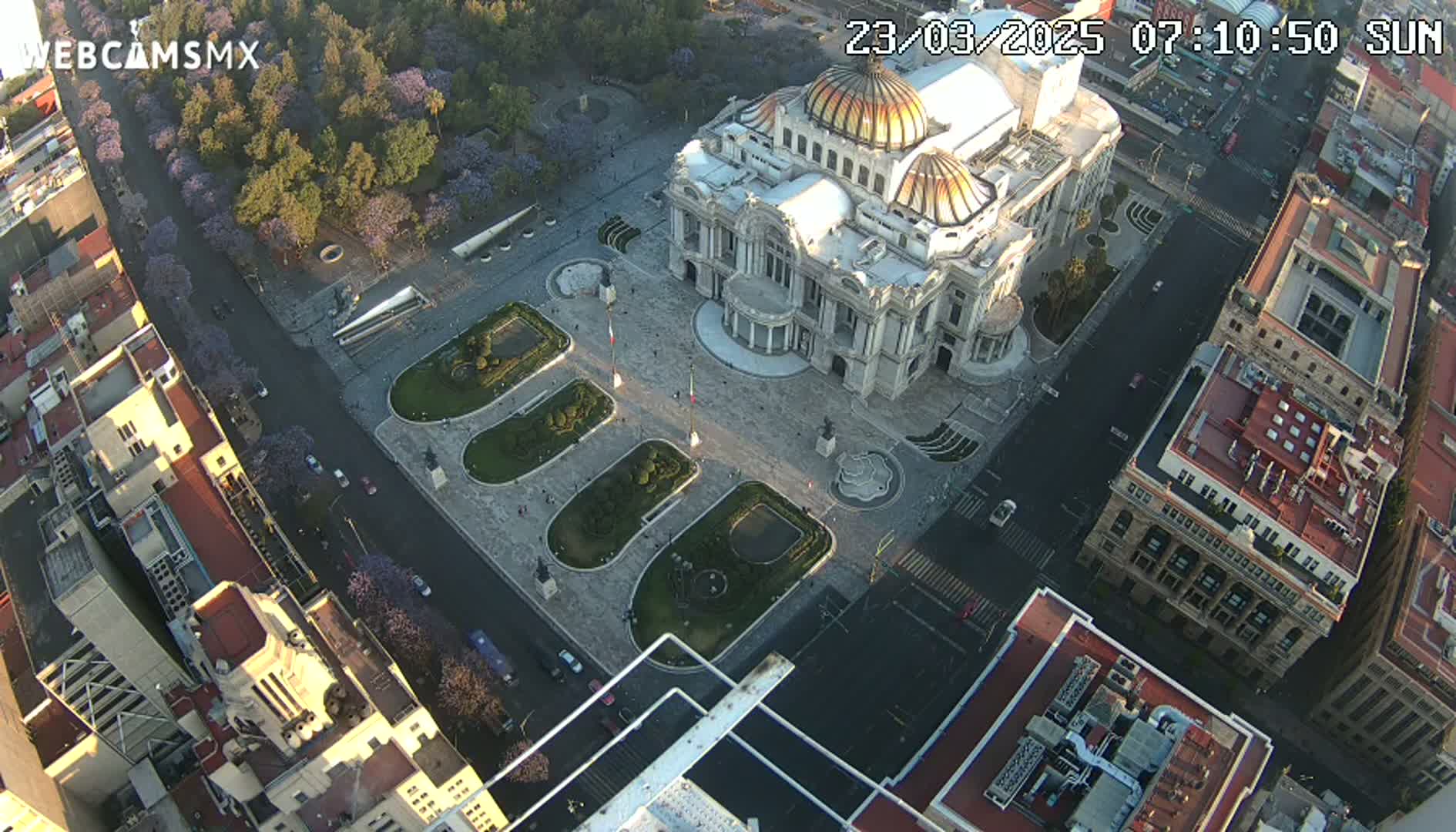 An aerial view shows a large, ornate building surrounded by a plaza with landscaped areas and city streets, under a sunny sky.