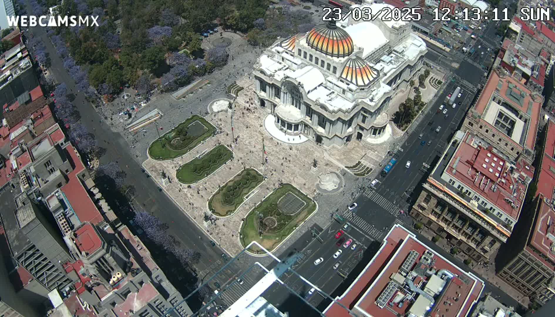 An aerial view shows a large plaza with a grand building in the center, surrounded by city streets and buildings on a sunny day.