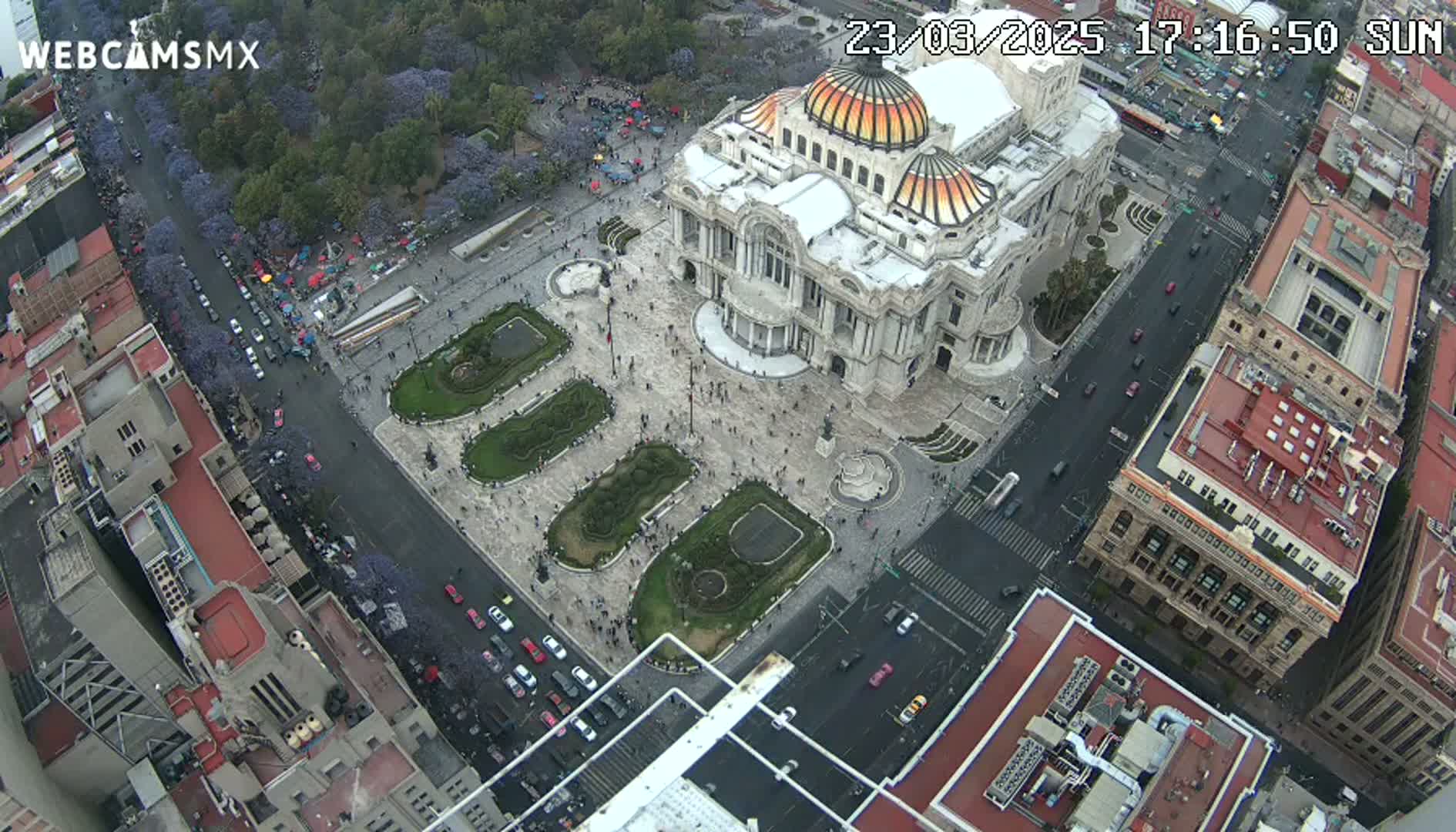 An aerial view shows a large, ornate building in a city square surrounded by streets and other buildings on a sunny day.