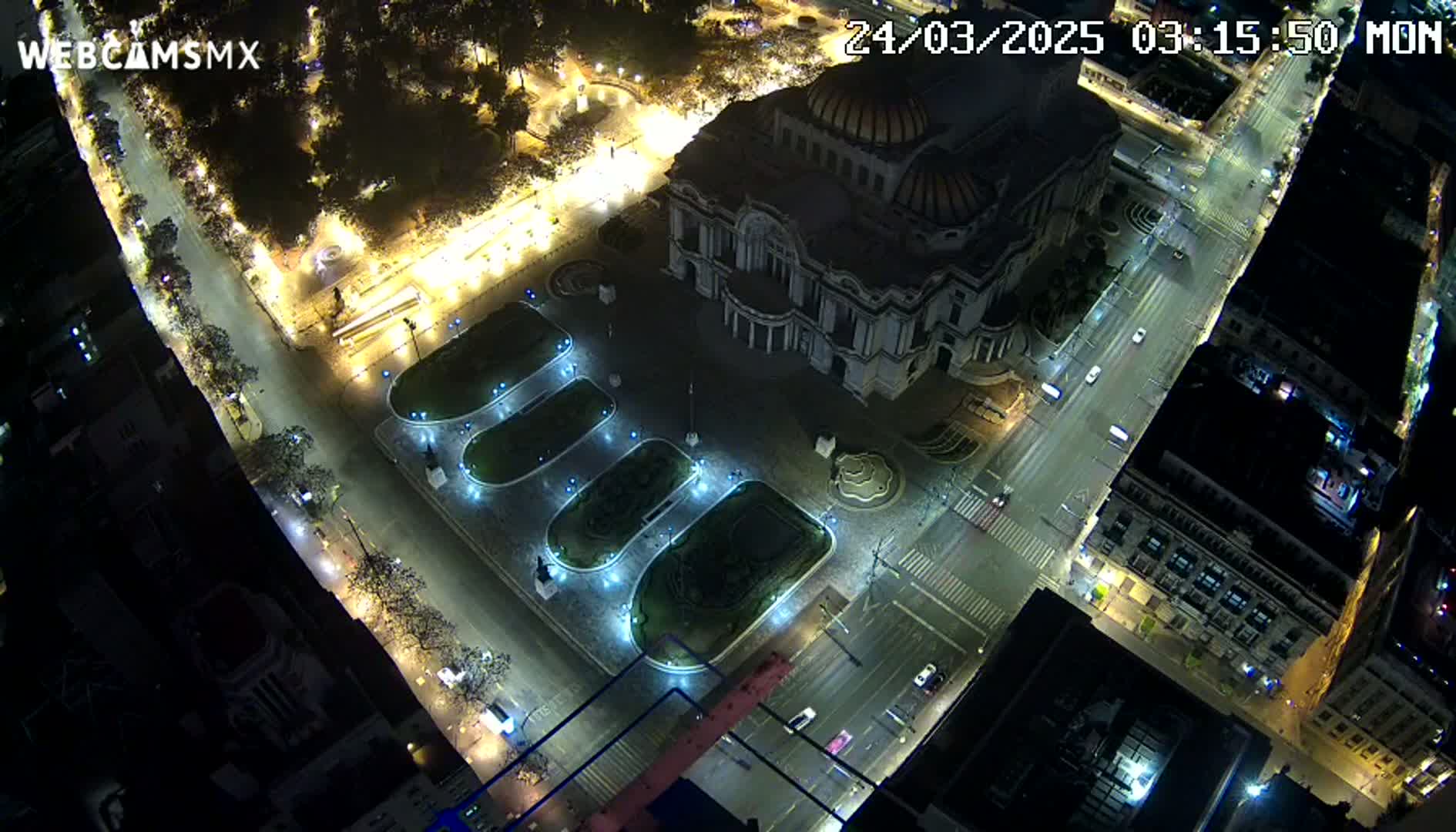 A nighttime aerial view shows a large, illuminated building surrounded by streets and a plaza with landscaped areas, lightly trafficked with cars.