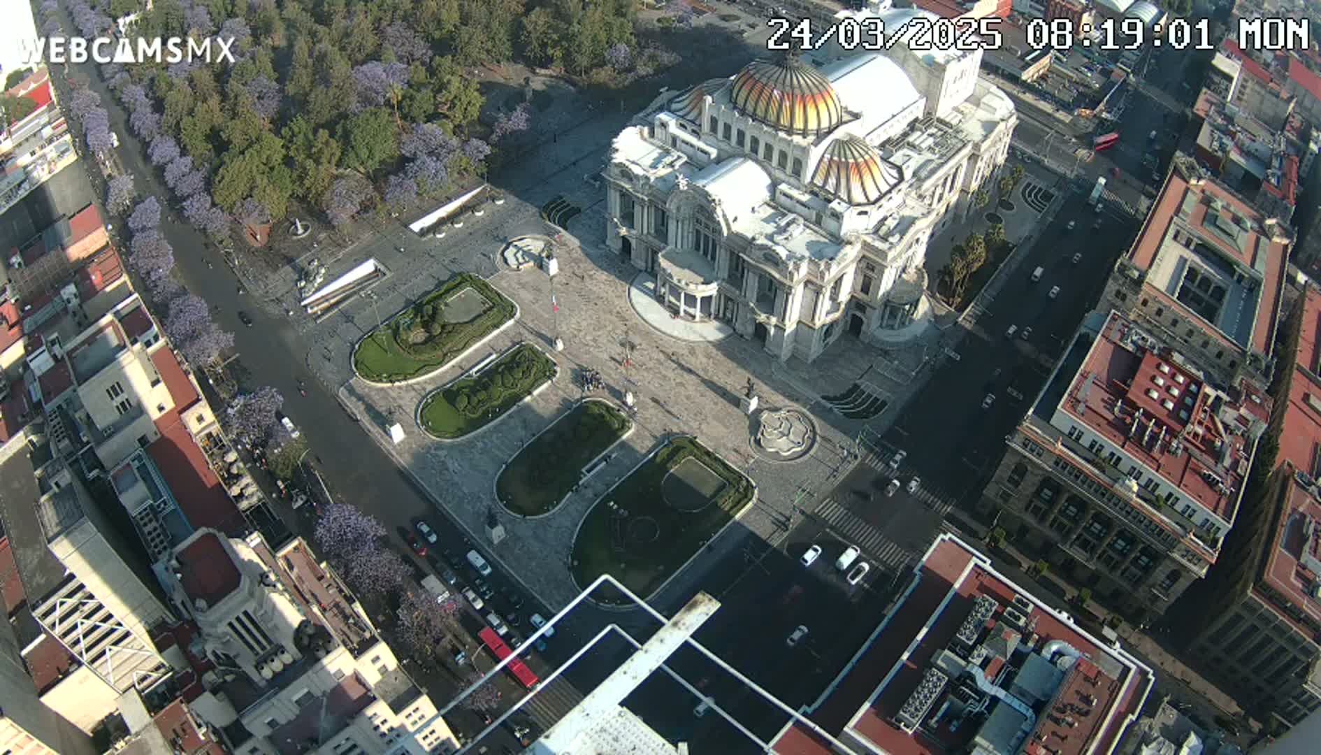 An aerial view shows a large, ornate building surrounded by a plaza with landscaped gardens, streets, and other buildings, under sunny conditions.