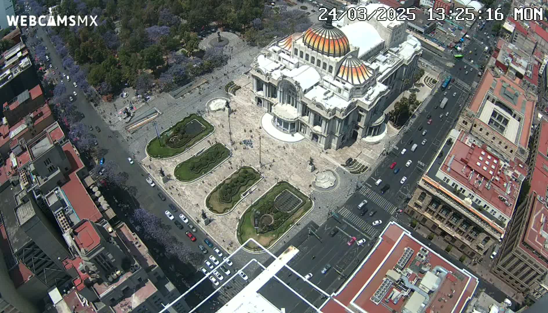 An aerial view shows a large palace-like building in a city square surrounded by streets with cars and buildings, under a clear sky.