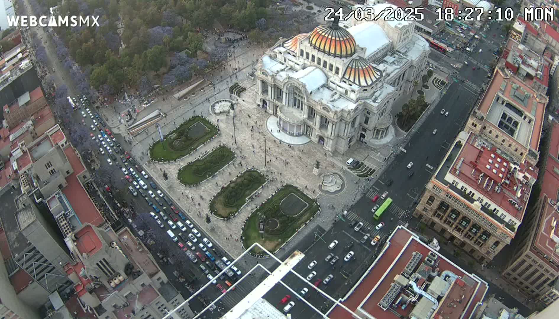 An aerial view shows a large, ornate building surrounded by a plaza with landscaped gardens and busy city streets filled with vehicles under a mostly clear sky.