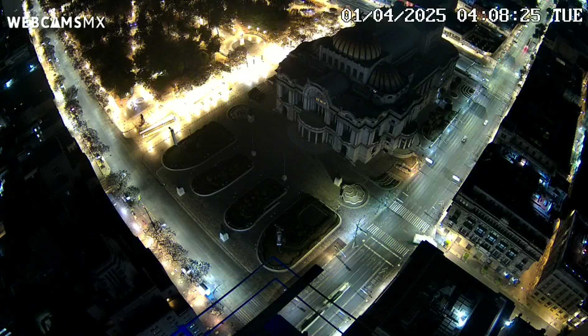 An aerial nighttime view of a city square with a large building, illuminated streets, and sparse traffic.