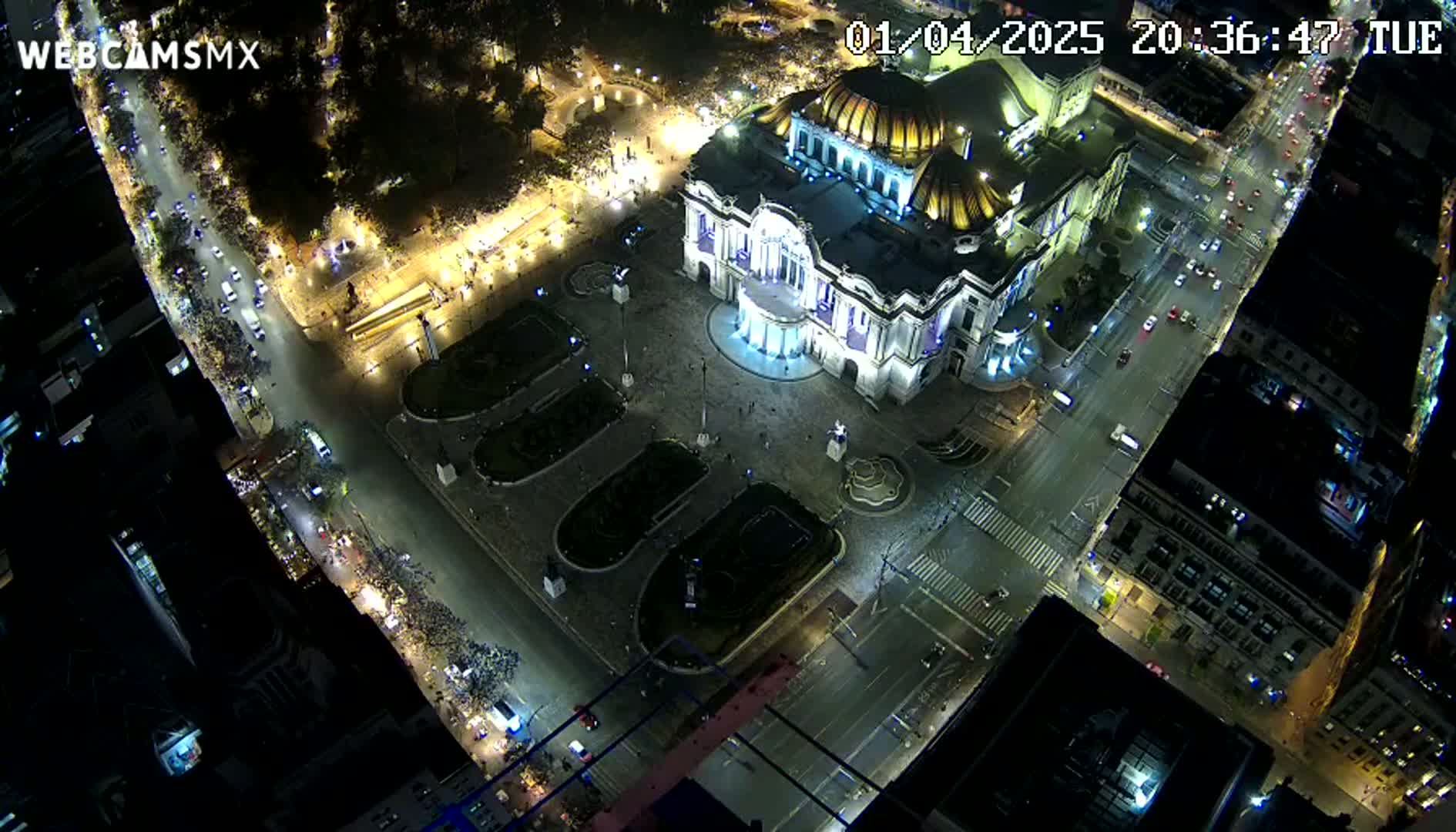 A nighttime aerial view shows a large, illuminated building surrounded by streets with cars and a crowd of people gathered in a plaza.