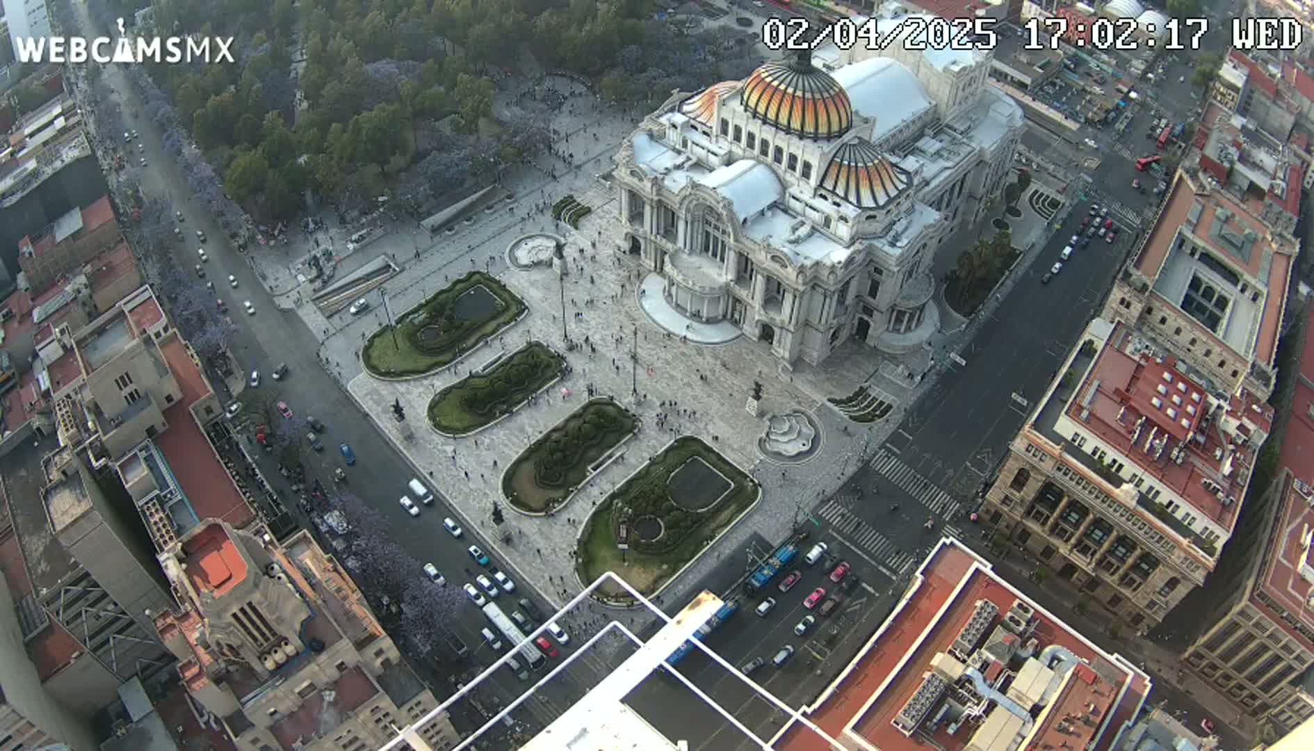 An aerial view shows a large building with a dome surrounded by a plaza with landscaped gardens and city streets with cars under a clear sky.