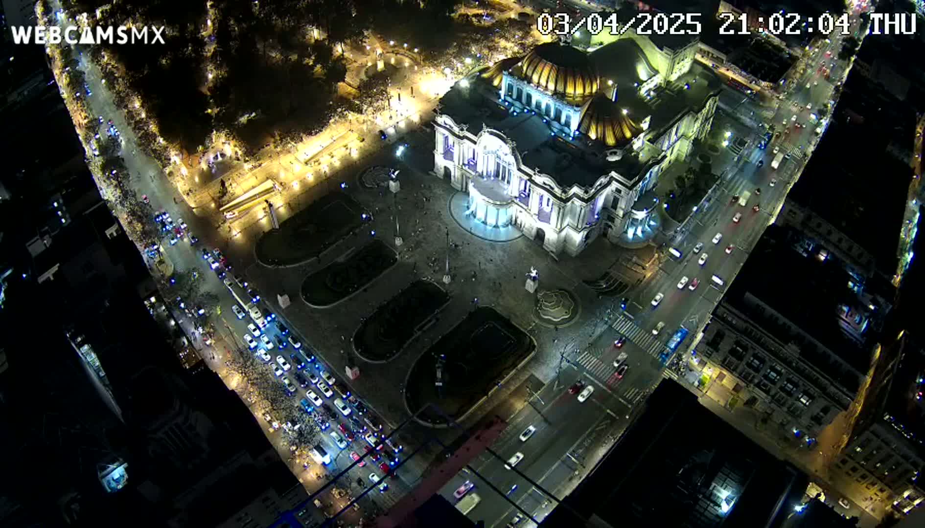 An aerial nighttime view shows a large building illuminated, surrounded by streets with cars and a plaza, under a clear, dark sky.