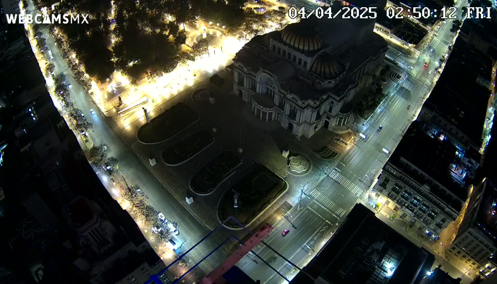 A nighttime aerial view shows a large, illuminated building surrounded by city streets with sparse traffic, and a plaza with landscaping.