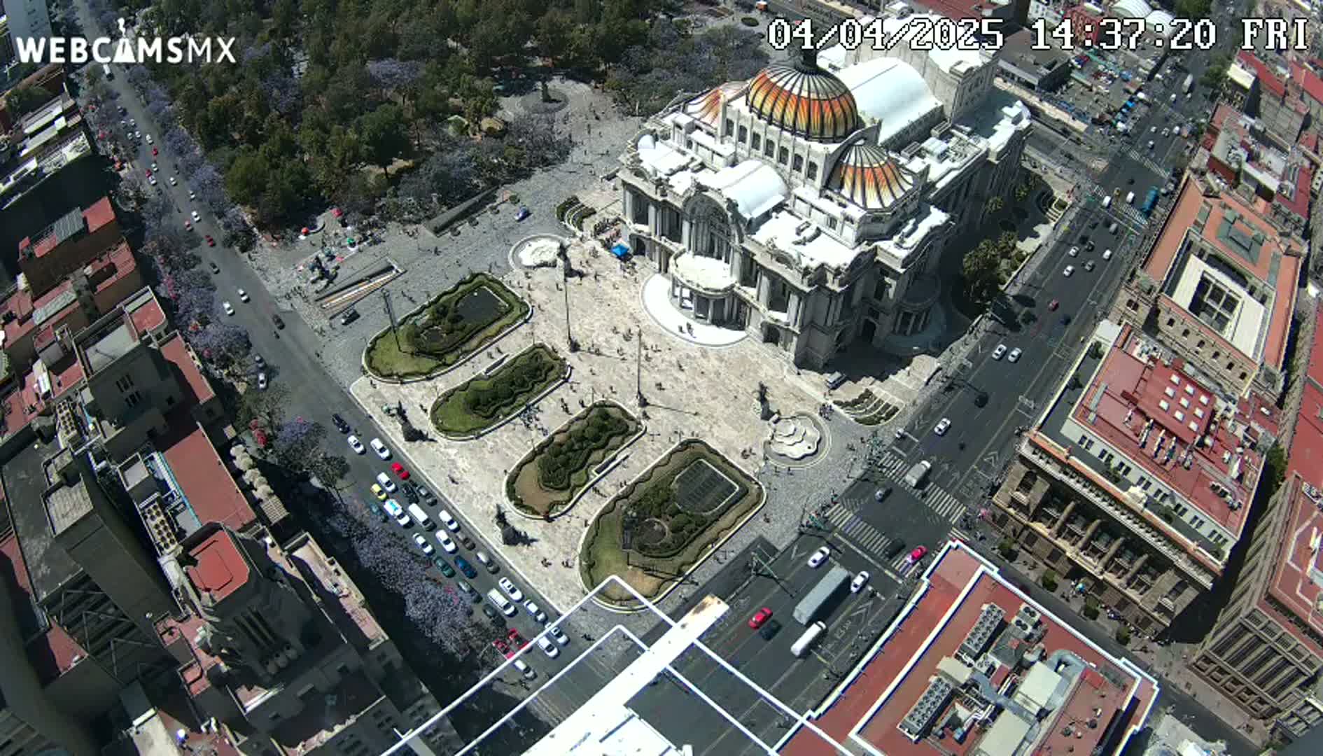 An aerial view shows a large, ornate building in a city square surrounded by streets and other buildings on a sunny day.