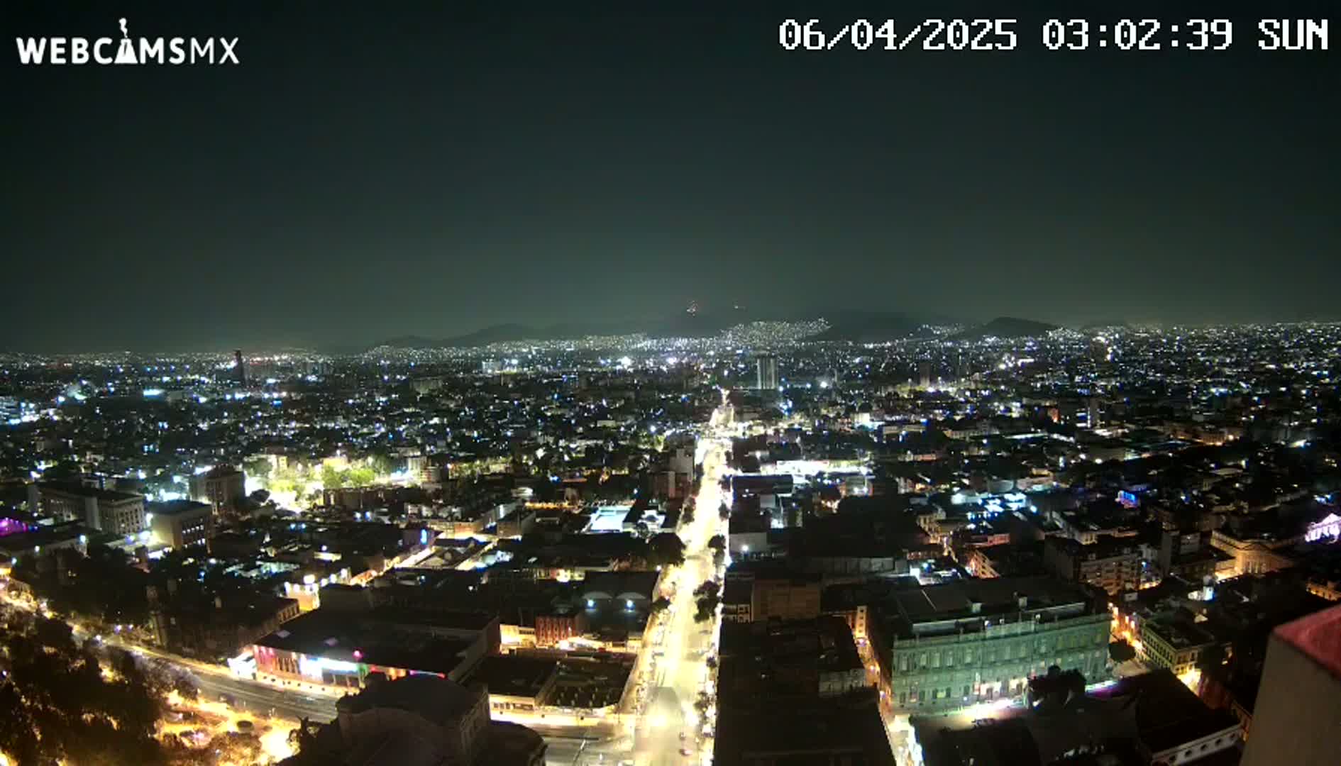 A nighttime, aerial view of a sprawling city illuminated by countless lights, with distant mountains visible under a clear sky.