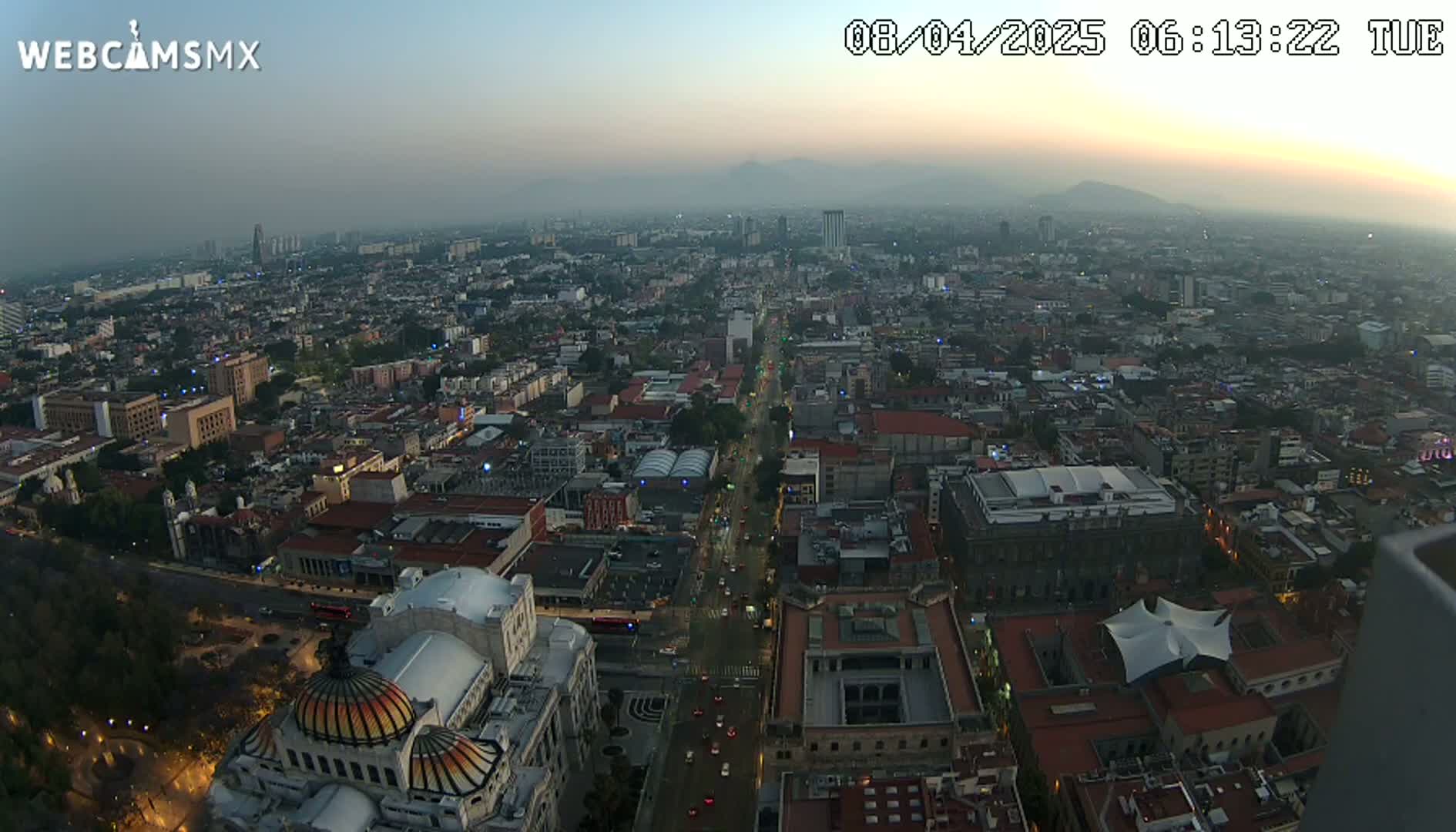An aerial view of a large city at dusk under hazy conditions, showing a long street with traffic and many buildings, including a large dome-shaped structure.